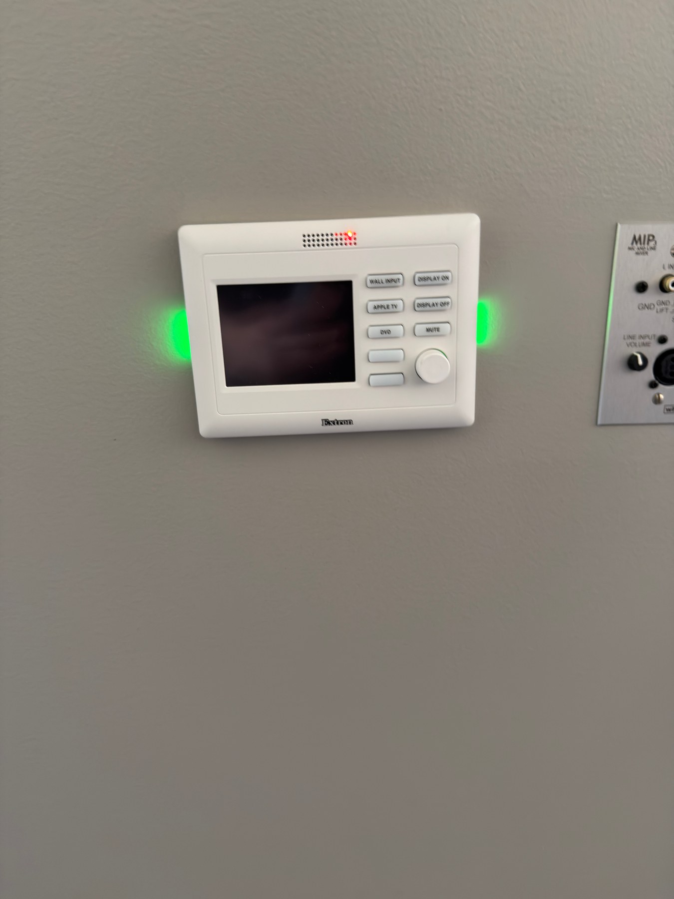 Previous wall-mounted Extron controller with a small screen, fixed source buttons, volume knob, and illuminated side indicators